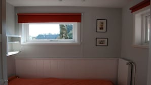 Ein Zimmer mit einem Einzelbett, einem Fenster mit rotem Vorhang, einem Heizkörper und zwei gerahmten Bildern an der Wand.