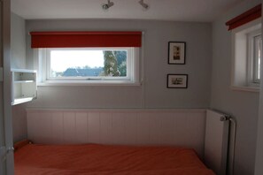 Une chambre avec un lit simple, une fenêtre avec un store rouge, un radiateur et deux tableaux encadrés accrochés au mur.