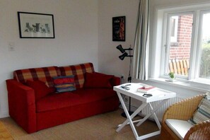 Un canapé rouge avec un plaid, une table blanche avec une télécommande et un fauteuil en osier.