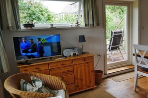 Ein Wohnzimmer mit Fernseher, Holzschrank, Korbstuhl und Blick ins Freie durch eine Schiebetür.