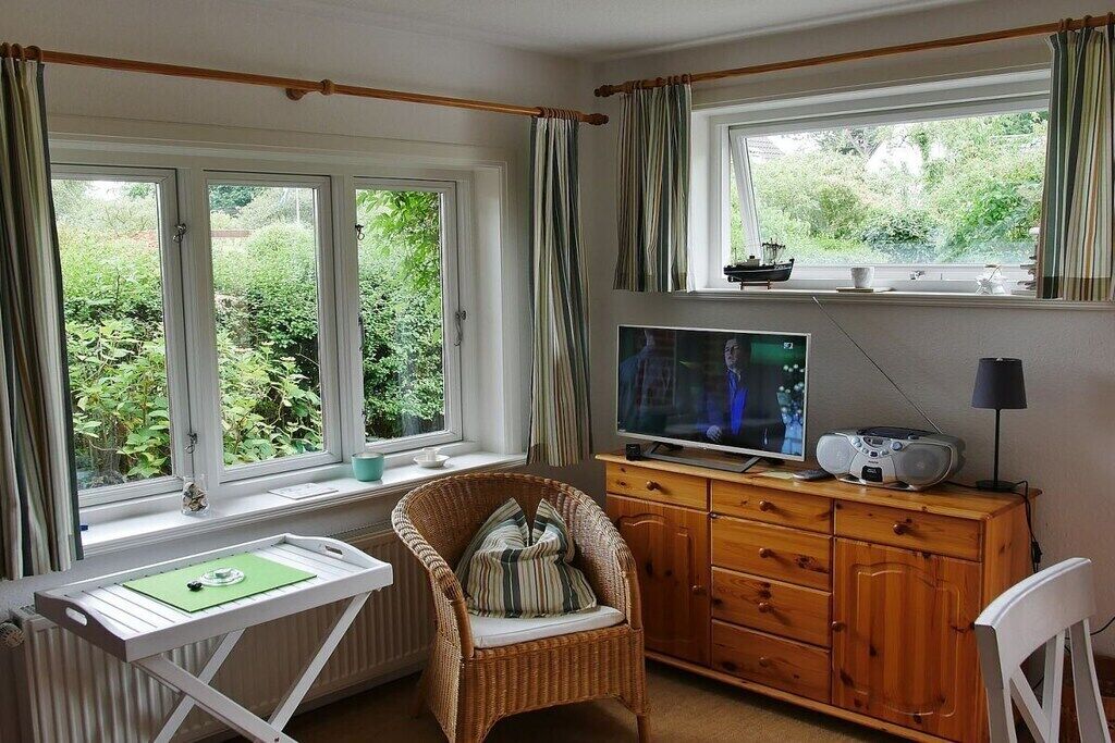 Quarto com televisão, cadeira de vime, armário de madeira e janela com cortinas.