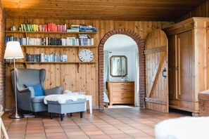 A cozy room with a wooden bookshelf, a comfortable armchair, a small table, and a large arched doorway leading to another room. - Terrace house by the sea (Langballig)