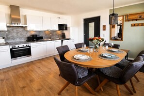 Une cuisine moderne avec des armoires blanches, un four encastré et un coin repas doté d’une table en bois, mise pour six personnes.