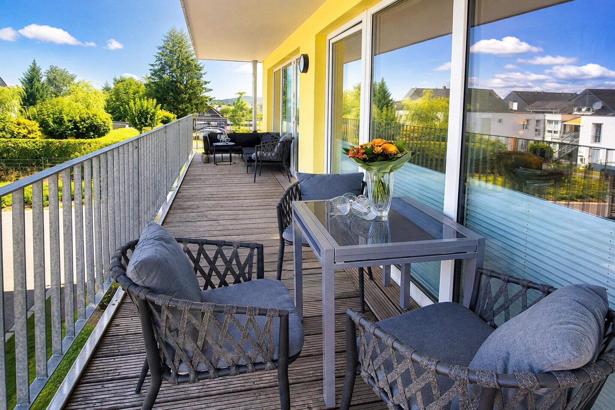 A balcony with a table and chairs, a vase with flowers, and a view of the neighborhood.