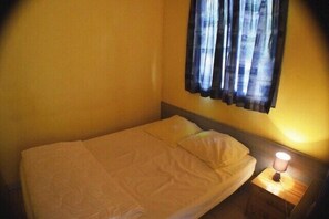 A single bed with a white bedspread and pillows, a nightstand with a lamp, and a window with curtains. - Chalet #14, Sleeps 4, Domaine de Ribière (Grospierres)