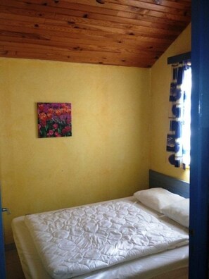 A single bed with a white mattress and pillows, a wooden ceiling, a yellow wall, and a colorful painting. - Chalet #15, sleeps 4, Domaine de Ribière (Grospierres)
