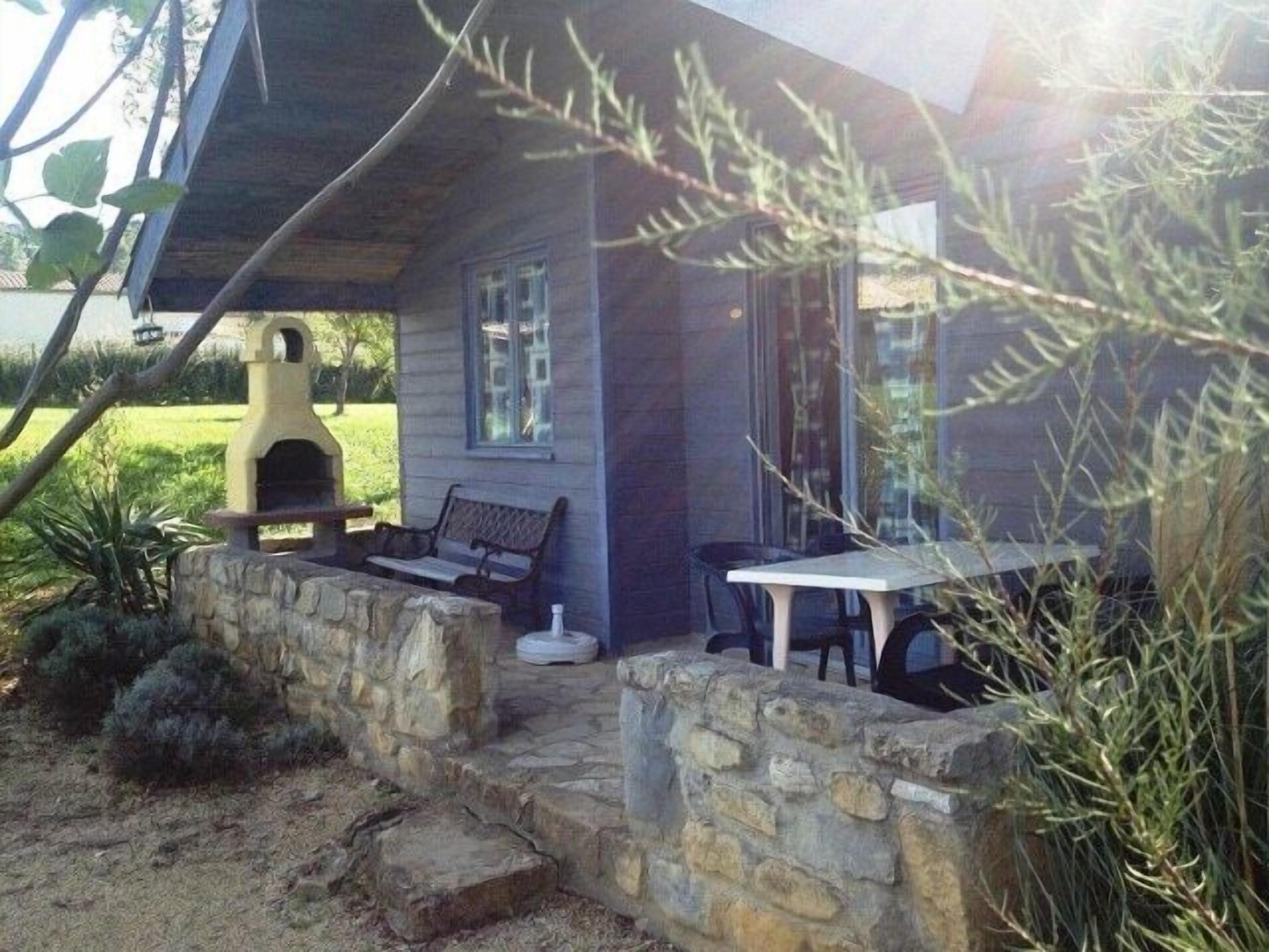 A stone-walled outdoor seating area with a bench and table, a barbecue grill, and a house with a window.