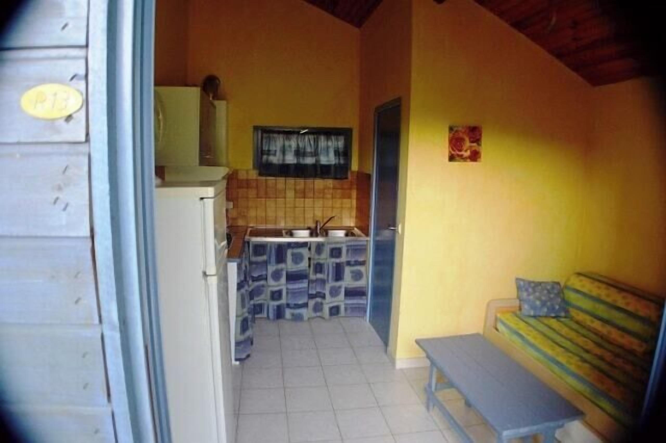 A compact kitchen with a sink, a small refrigerator, and a window above the counter. There is a bench with a cushion in the adjacent room.
