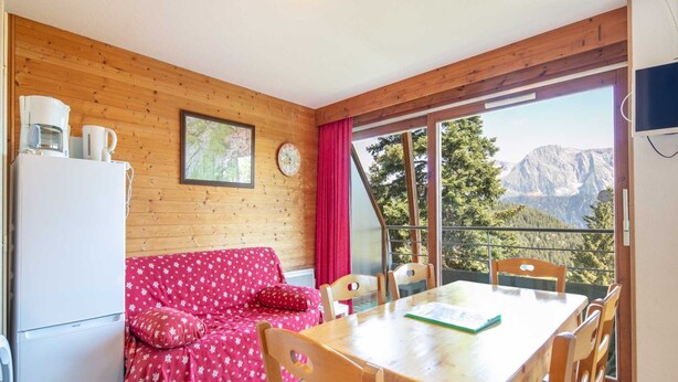 A cozy room with a wooden interior, a dining table with chairs, a red sofa, a white refrigerator, and a window with a mountain view. - Alpine Ski Apartment in Tignes for 4 (Chamrousse)