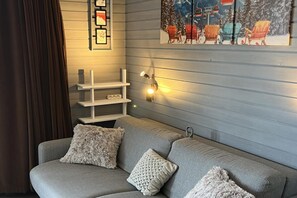 A modern living room with a grey sofa, decorative pillows, wall-mounted shelves, and a painting of winter scene. - Alpine Ski Apartment in Tignes for 4 (Orcières)