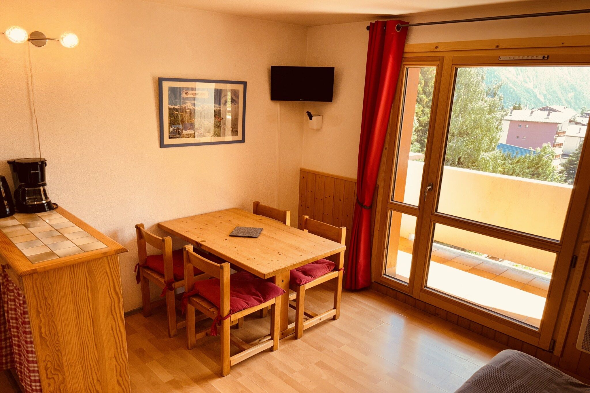 A room with a wooden table and chairs, a television mounted on the wall, and a window with a view of buildings.