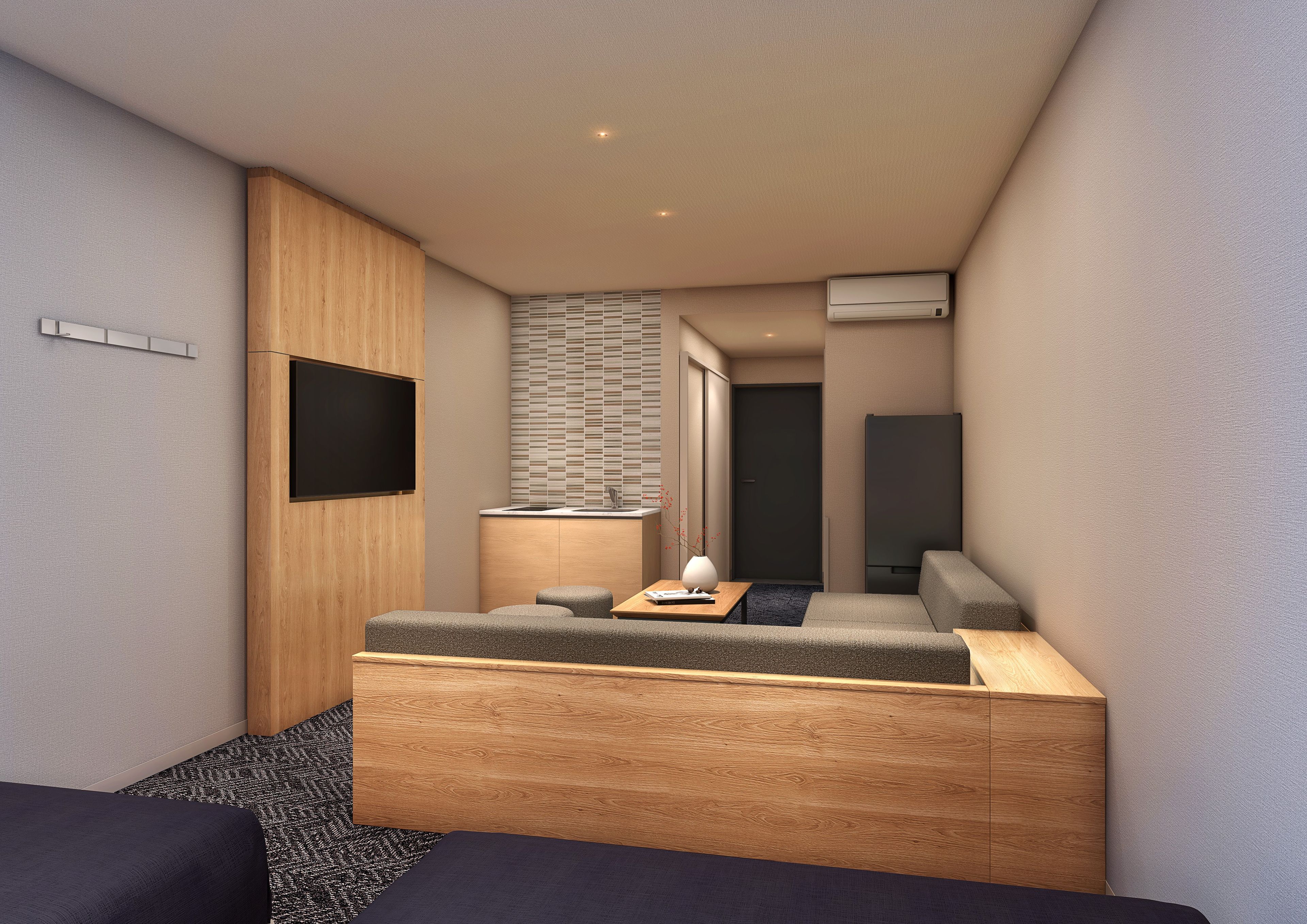 A compact hotel room with a sofa bed, a small table with a lamp, a TV mounted on the wall, a window with blinds, and an air conditioning unit.