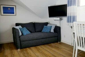 A living room with a dark grey sofa, a flat-screen TV mounted on the wall, and a white crib. - Holiday home with 2 bedrooms (Wangerland Hooksiel)