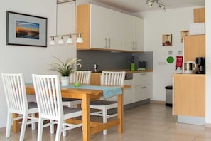 A modern kitchen with white cabinets, a wooden dining table with chairs, and a small island with a microwave. - Holiday home with 2 bedrooms (Wangerland Hooksiel)
