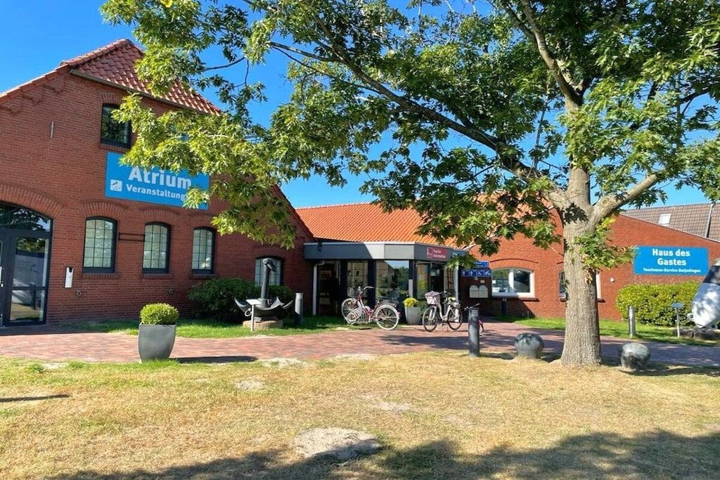 Un bâtiment avec une enseigne indiquant « Atrium Verwaltung » et une autre enseigne indiquant « Haus des Gastes ». On peut voir des vélos garés à l’extérieur, un arbre au premier plan et un chemin piétonnier aménagé.