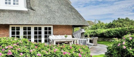 Una casa con techo de paja, fachada de ladrillo y un jardín con flores rosadas.