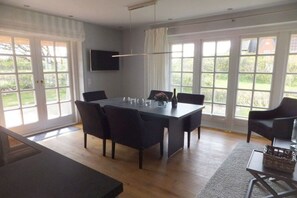 Un comedor con una mesa y sillas, un televisor montado en la pared y ventanas grandes con cortinas.