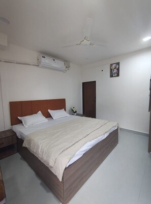 Quarto de hotel com cama, mesa de cabeceira, ar-condicionado fixado na parede, porta e ventilador de teto.