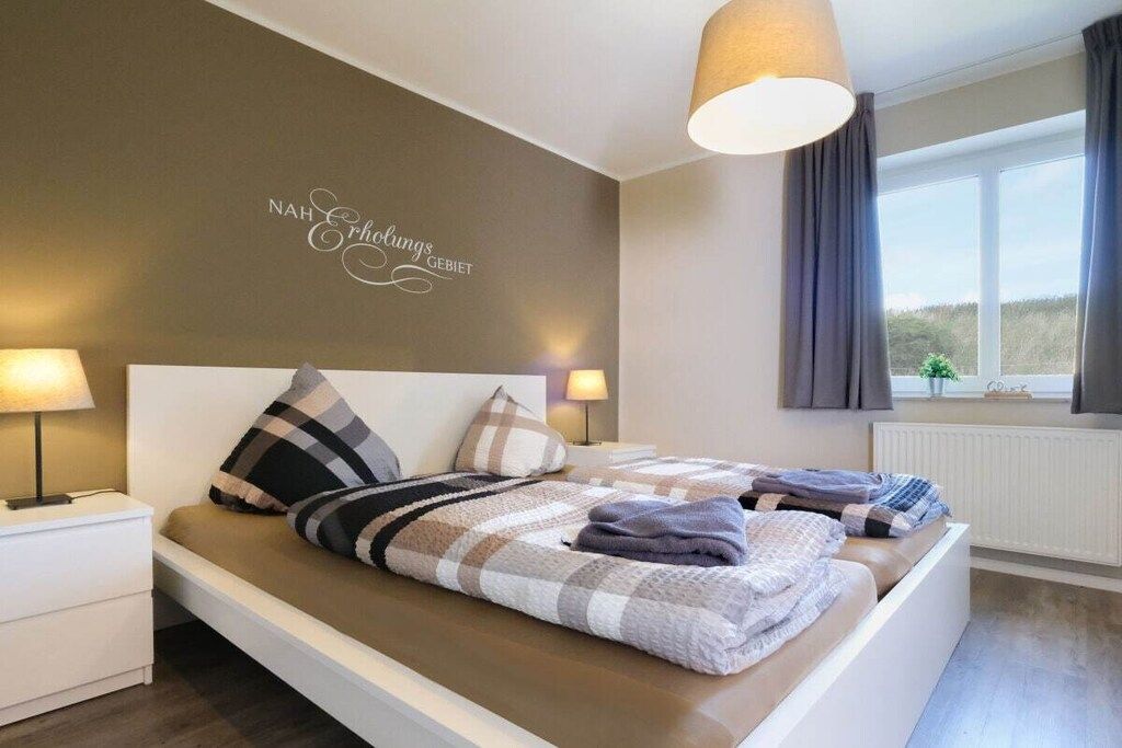 Une chambre à coucher comprenant un lit, des tables de chevet, une lampe fixée au mur, une fenêtre avec des rideaux et une vue sur l’extérieur.
