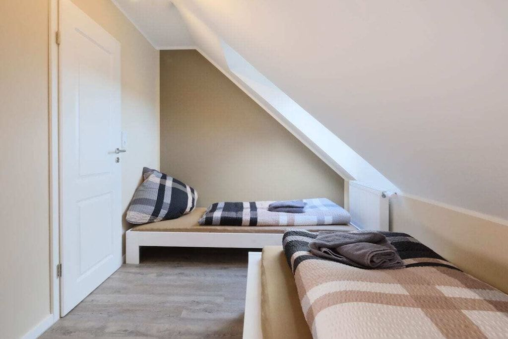 A small, cozy room with a sloped ceiling, a bed with a checkered bedspread, a bench with pillows, and a white door.