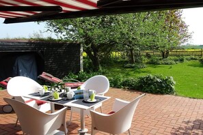 An outdoor patio with a table set for two, surrounded by white chairs, a covered area with a red and white striped awning, and a view of a lush garden and a wooden fence. - Holiday home in Schlei with 3 bedrooms (Kappeln Stadt)