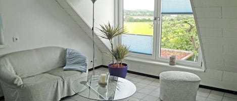 Una habitación con un sofá, una pequeña mesa redonda, una planta en maceta y vistas a un paisaje verde a través de una ventana.