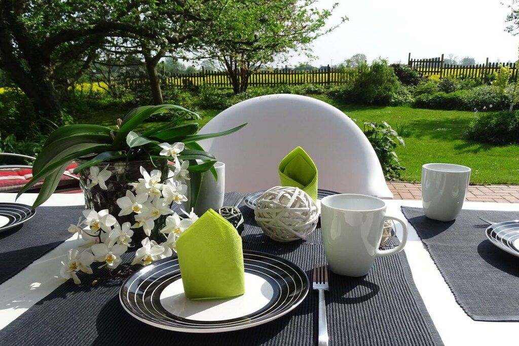 Disposición de comedor al aire libre con una mesa preparada para dos personas, con platos blancos, servilletas verdes y vasos blancos.