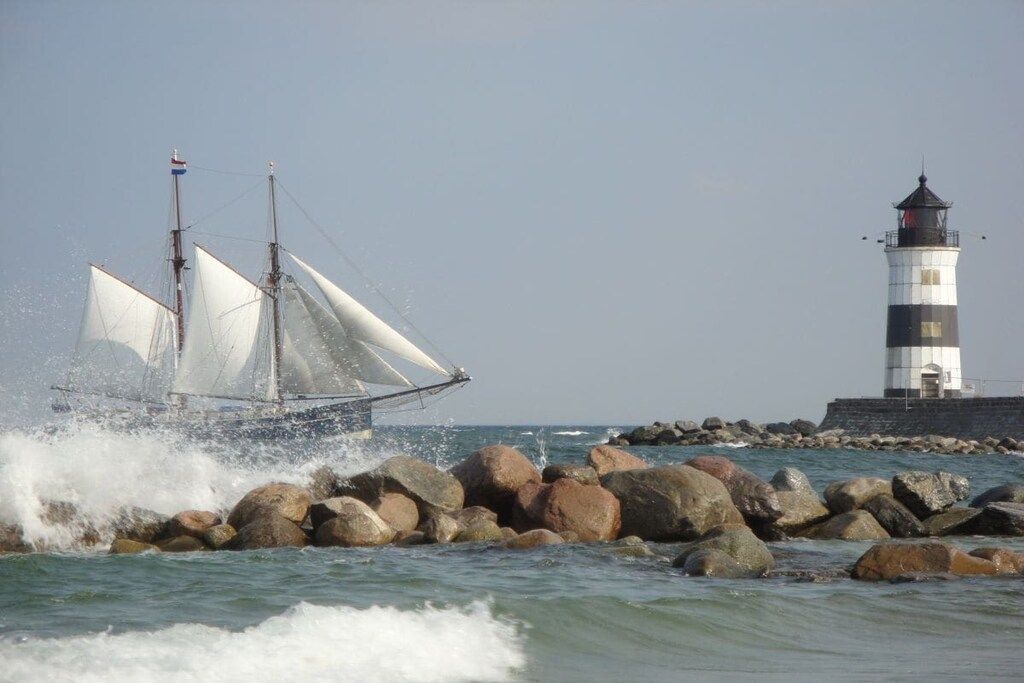 Ein Segelboot mit weißen Segeln steuert in der Nähe eines Leuchtturms und einer felsigen Küste.