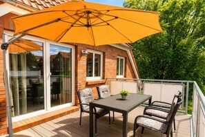 A deck with a table and chairs, an umbrella, and a brick house with glass doors. - Holiday apartment with 2 bedrooms (Carolinensiel)