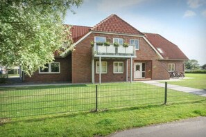 A two-story brick house with a red-tiled roof, a balcony with plants, and a fenced yard. - Holiday apartment with 2 bedrooms (Carolinensiel)