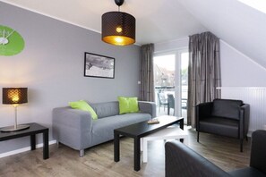 Una sala moderna con un sofá gris, un sillón negro y una mesa de centro. Hay una lámpara en una mesita auxiliar, un cuadro enmarcado en la pared y una ventana con cortinas.