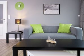 A modern living room with a grey sofa, two green cushions, a black coffee table, a small side table with a lamp, and a framed picture on the wall. - Holiday apartment with 2 bedrooms (Carolinensiel)