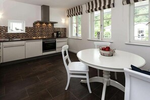 A modern kitchen with dark flooring, white cabinetry, a round dining table with two chairs, and a window with a view of greenery. - No. 2 (Veranda) in Villa Fabiola (Ostseebad Binz)