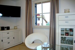 Ein Zimmer mit einem an der Wand befestigten Flachbildfernseher, einer weißen Kommode mit Schubladen, einem gewölbten weißen Sessel, einem Glastisch mit einer Tasse und einem Fenster mit Vorhängen.