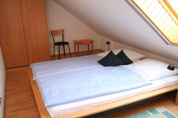 Una cama individual con estructura de madera, una mesita, una silla y un armario en una habitación con techos inclinados y un tragaluz.