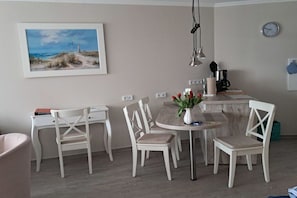 Un coin repas avec une table ronde, des chaises blanches, un vase avec des fleurs rouges, un tableau représentant une scène de plage et une horloge au mur.