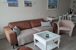 Ein Wohnzimmer mit einem braunen Sofa, einem Glastisch, einem weißen Beistelltisch und einem pinken Sessel. An der Wand hängen gerahmte Bilder.