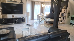 Uma sala de estar moderna com um sofá de couro preto, uma mesa de centro redonda, uma TV de tela plana e um espaço de jantar com uma mesa e cadeiras brancas.