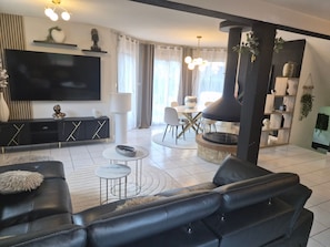 Un salon moderne avec un canapé en cuir noir, une table basse ronde, un téléviseur à écran plat et un coin repas comprenant une table et des chaises blanches.