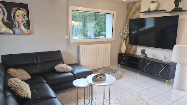 Uma sala de estar moderna com um sofá de couro preto, uma mesa de centro redonda, uma TV de tela plana e uma janela grande.