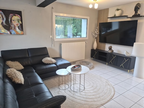 Un salon moderne avec un canapé en cuir noir, une table basse ronde, un téléviseur à écran plat et une grande fenêtre.