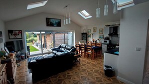 A spacious living area with a black leather sofa, a dining table with chairs, a kitchen area, and a large sliding glass door leading to a garden. - Bungalow Annex - Sleeps 6 With Countryside Views (Tarporley)