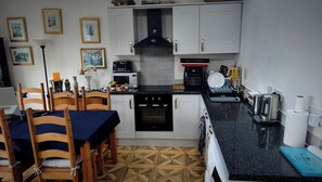 A compact kitchen with white cabinets, a black countertop, and a dining area with wooden chairs and a blue tablecloth. - Bungalow Annex - Sleeps 6 With Countryside Views (Tarporley)