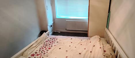 Une petite pièce étroite, avec un lit simple, un radiateur et une fenêtre équipée de stores.