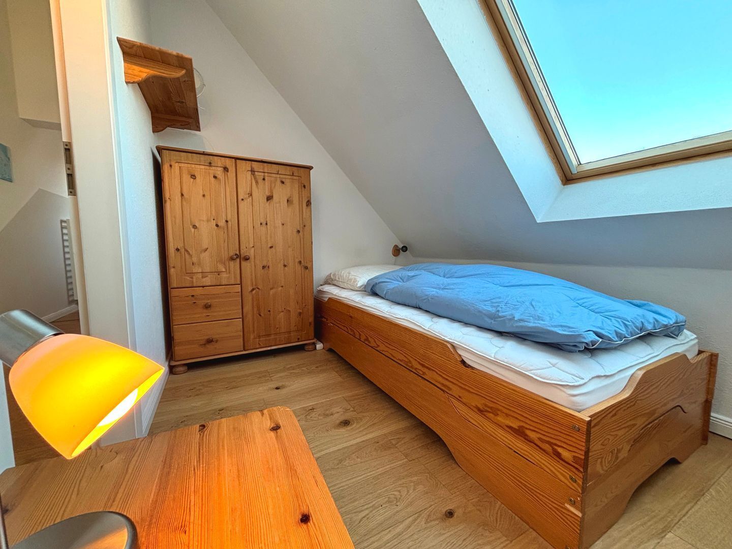 Una habitación pequeña e inclinada con una cama y un armario de madera, un escritorio con lámpara y un tragaluz.