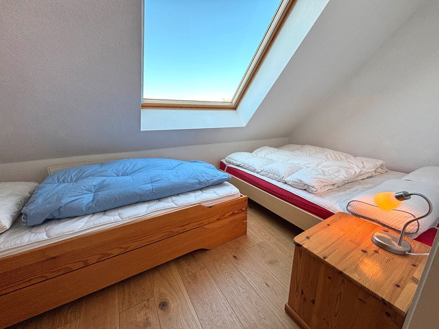 A small, compact room with two wooden beds, a skylight, and a bedside table with a lamp.