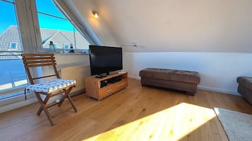 Una habitación con una televisión sobre un soporte de madera, un banco acolchado, una silla de madera y vistas de edificios a través de una ventana.