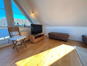 A room with a television on a wooden stand, a cushioned bench, a wooden chair, and a view of buildings through a window.