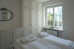 A bedroom with a bed, a radiator, a window with a view of greenery, and a wall with framed pictures. - Cozy Vacation Rental at Friesenring 17c in Wenningstedt (Wenningstedt-Braderup (Sylt))