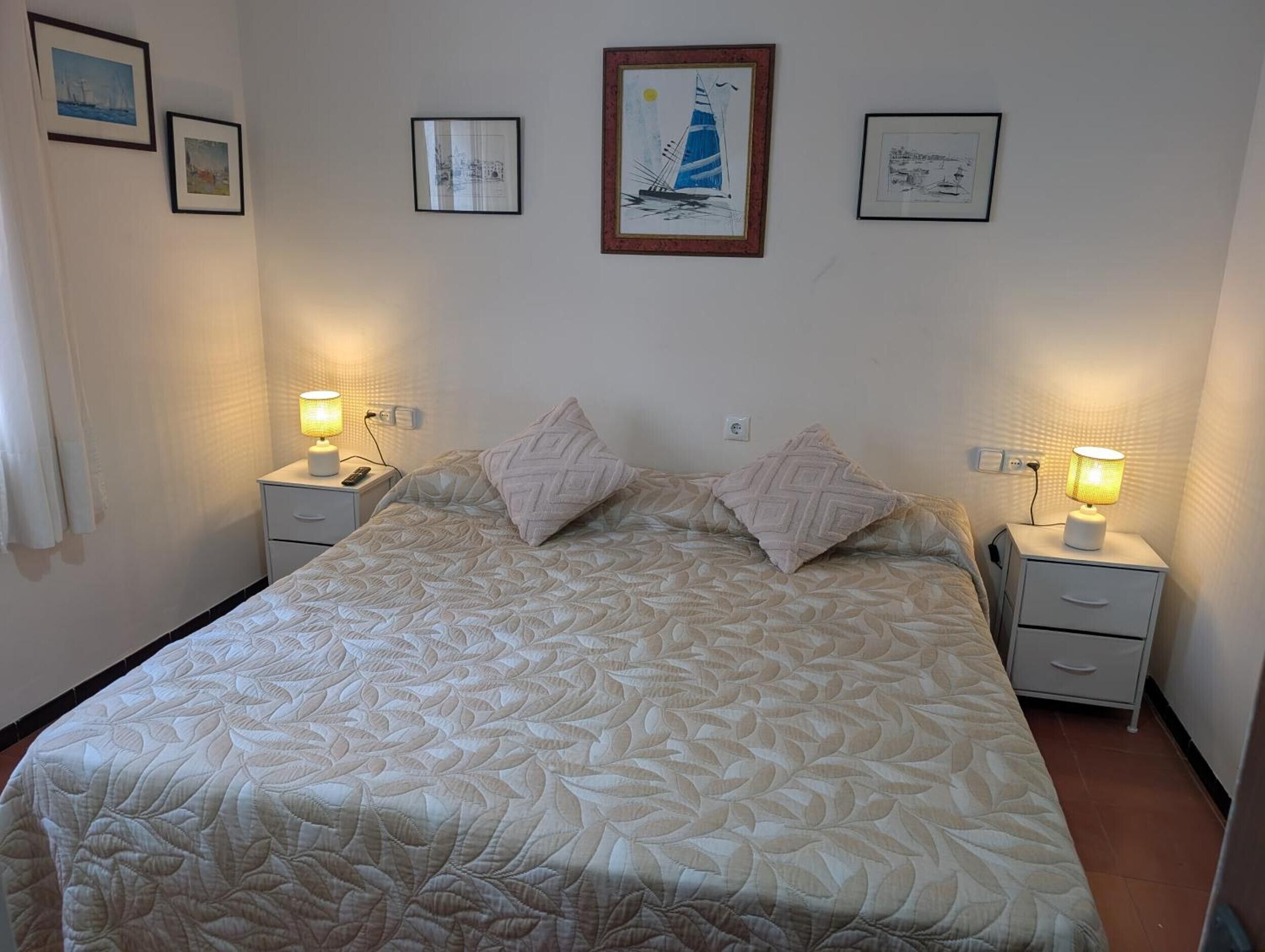 A bedroom with a bed, two bedside tables, a nightstand, and a wall with framed pictures.